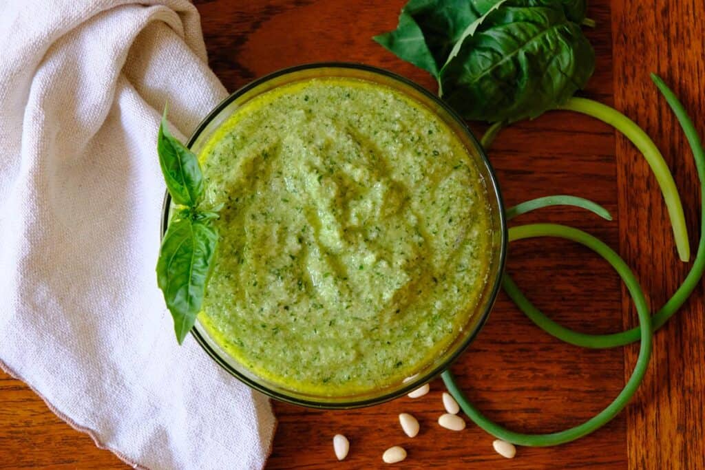 Glass bowl of homemade garlic scape pesto with fresh basil, pine nuts, garlic scapes, and leafy greens on a wooden table