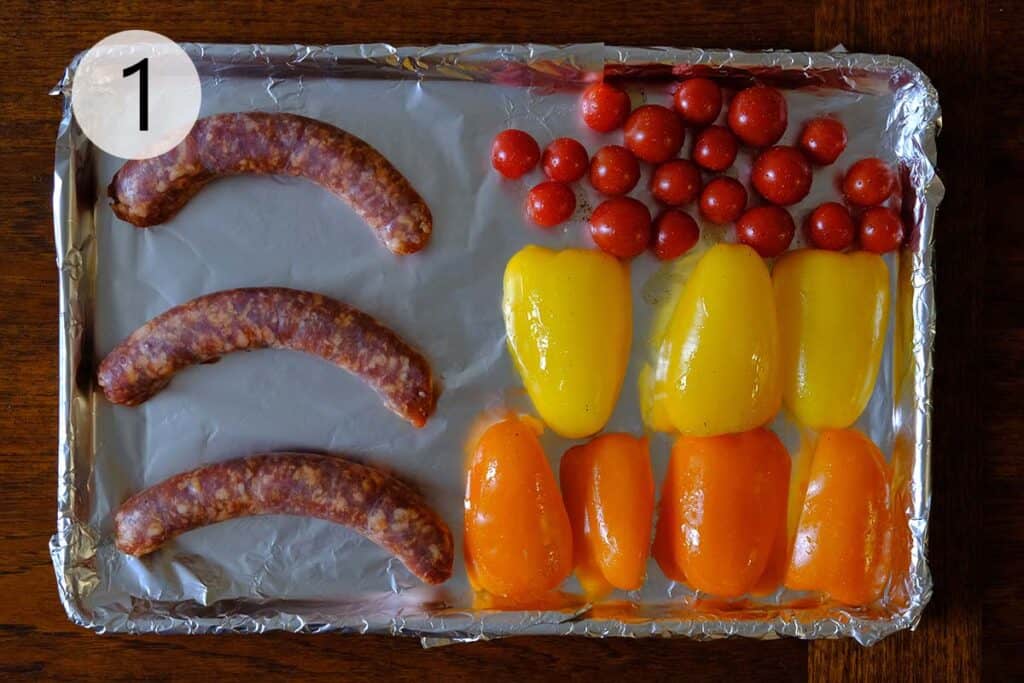 Sliced bell peppers and cherry tomatoes tossed with olive oil on a foil-lined baking sheet, ready to be roasted alongside Italian sausages.