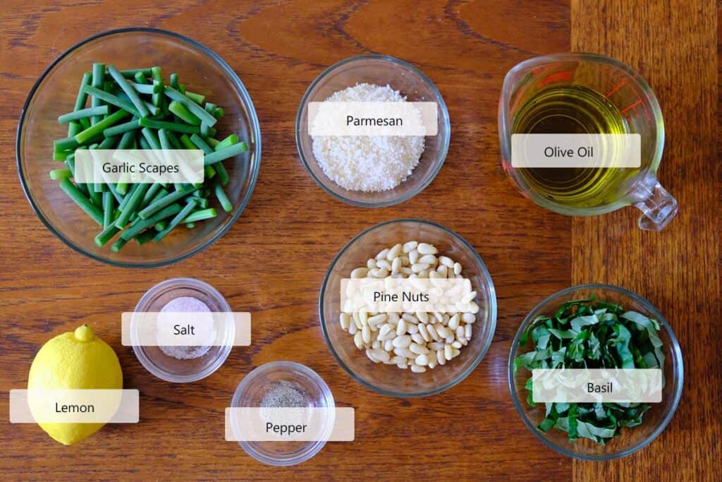 Garlic scape pesto ingredients arranged on a wooden surface, including garlic scapes, basil leaves, pine nuts, parmesan cheese, lemon, and olive oil