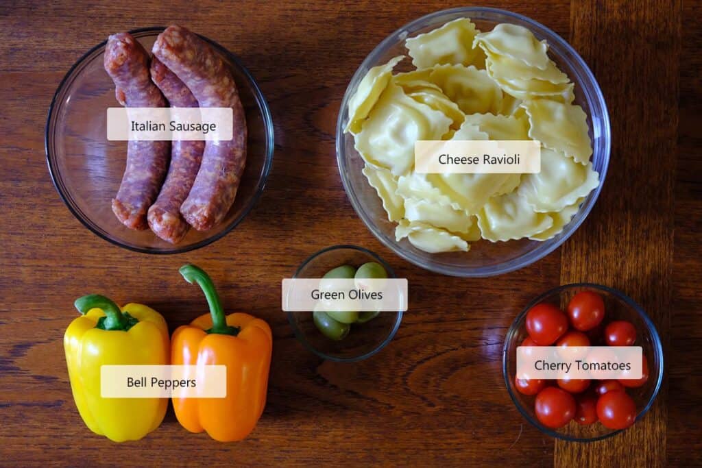 Flat lay of fresh ingredients for sausage pasta salad, including Italian sausage, bell peppers, cherry tomatoes, cheese ravioli, green olives, and olive oil—perfect for making an easy cold pasta salad with homemade dressing.