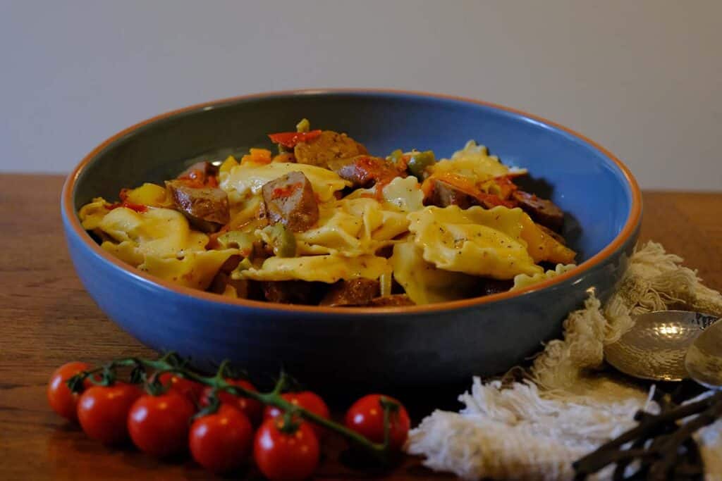 Large ceramic bowl filled with sausage pasta salad made with cheese ravioli, roasted peppers, cherry tomatoes, and sliced Italian sausage, served with fresh cherry tomatoes on the vine.
