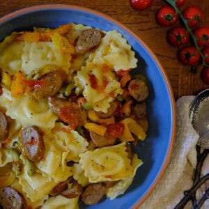 Large ceramic bowl filled with sausage pasta salad made with cheese ravioli, roasted peppers, cherry tomatoes, and sliced Italian sausage, served with fresh cherry tomatoes on the vine.