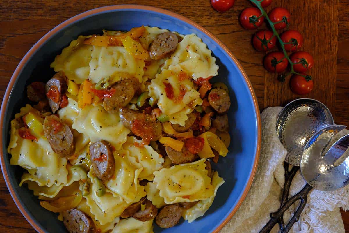 Large ceramic bowl filled with sausage pasta salad made with cheese ravioli, roasted peppers, cherry tomatoes, and sliced Italian sausage, served with fresh cherry tomatoes on the vine.