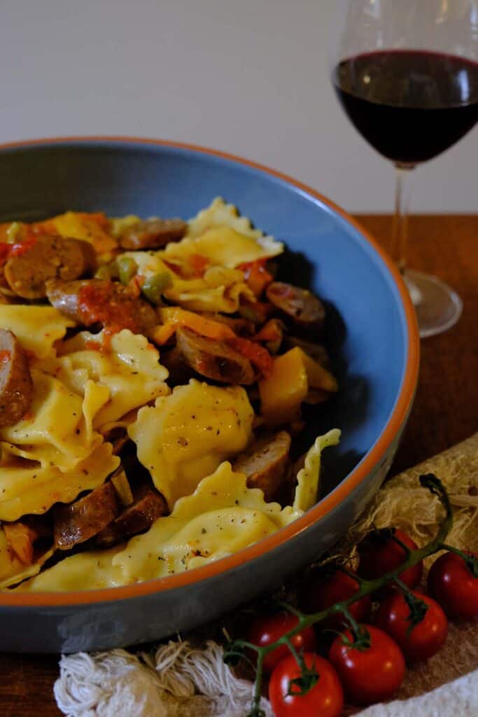 Large ceramic bowl filled with sausage pasta salad made with cheese ravioli, roasted peppers, cherry tomatoes, and sliced Italian sausage, served with a glass of red wine and fresh cherry tomatoes on the vine.