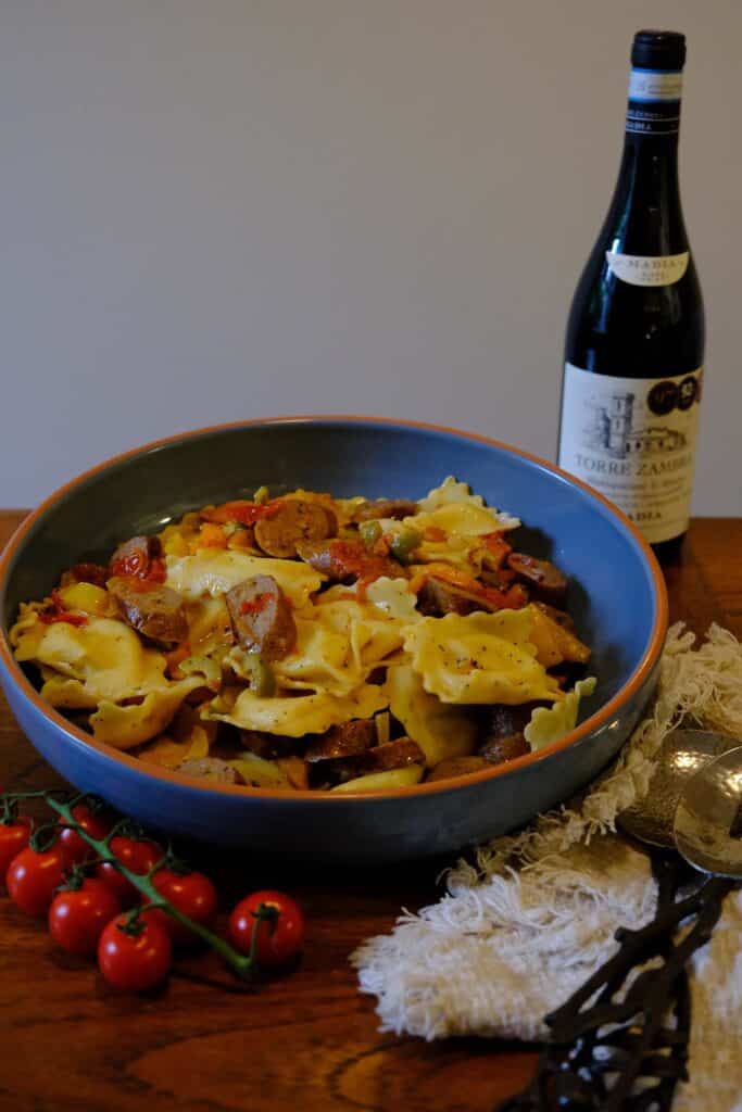 Large ceramic bowl filled with sausage pasta salad made with cheese ravioli, roasted peppers, cherry tomatoes, and sliced Italian sausage, served with a bottle of red wine and fresh cherry tomatoes on the vine.