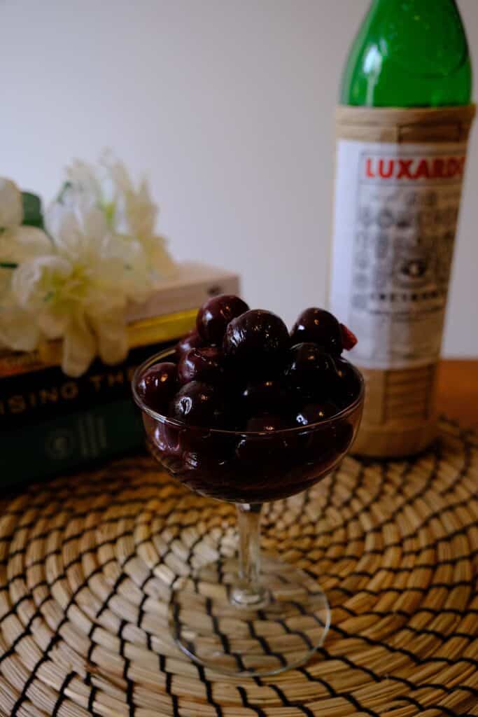 Easy Luxardo cherry recipe styled in a coupe glass with a bottle of Luxardo maraschino in the background on a woven placemat.