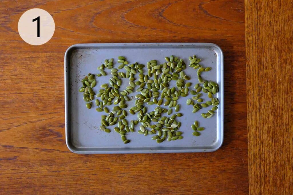 Close-up of pumpkin seeds tossed in olive oil and seasoning, spread on a baking sheet on a wooden surface.