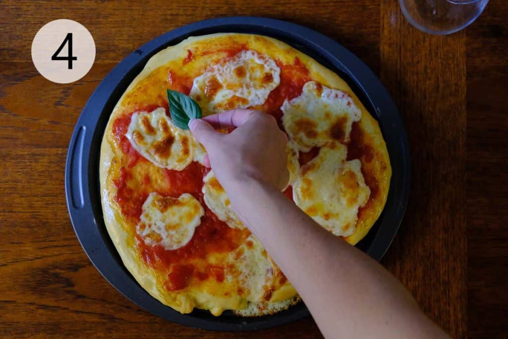 Golden brown Margherita pizza fresh out of the oven with bubbly mozzarella and charred crust with fresh basil leaves being placed on top.
