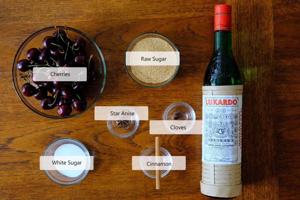 Ingredients for homemade Luxardo cherry recipe including fresh cherries, raw sugar, white sugar, whole spices, and Luxardo maraschino liqueur arranged on a wooden table