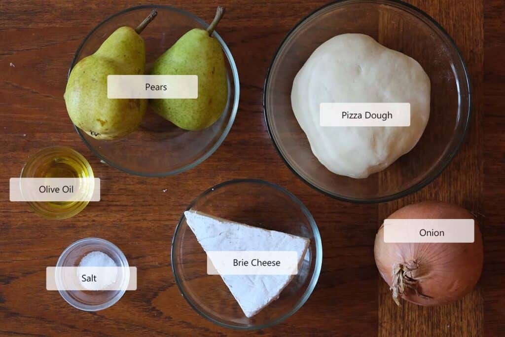 Overhead shot of fresh ingredients for pear pizza recipe including pears, pizza dough, onion, brie cheese, olive oil, and salt – easy and gourmet pizza prep.