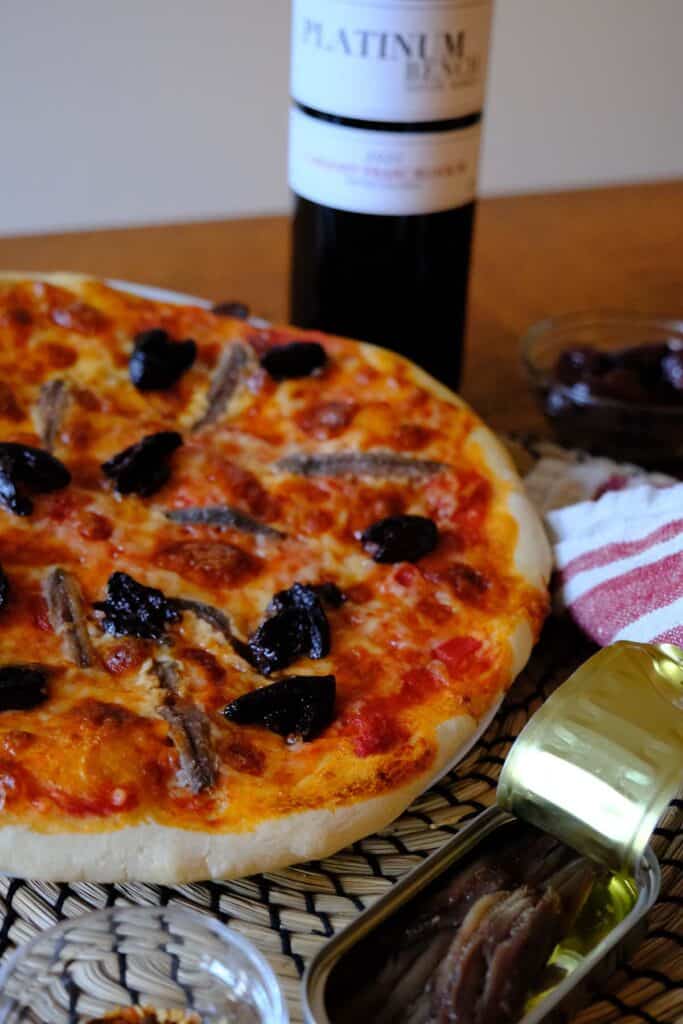 Close-up of a baked anchovy pizza with black olives and mozzarella, surrounded by anchovy fillets and a bottle of wine.