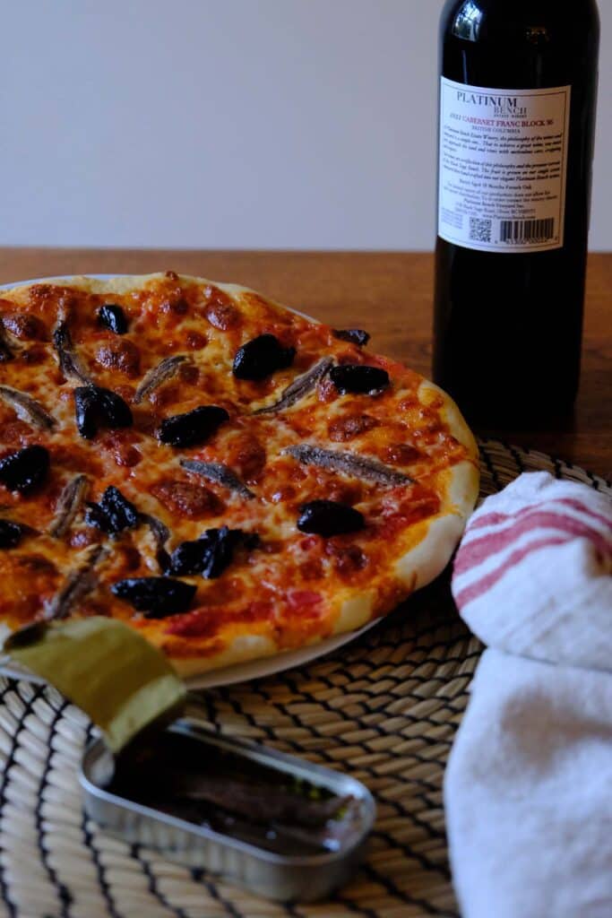 Homemade anchovy pizza recipe with melted cheese and olives on a plate, styled with anchovy can, wine, and kitchen towel.