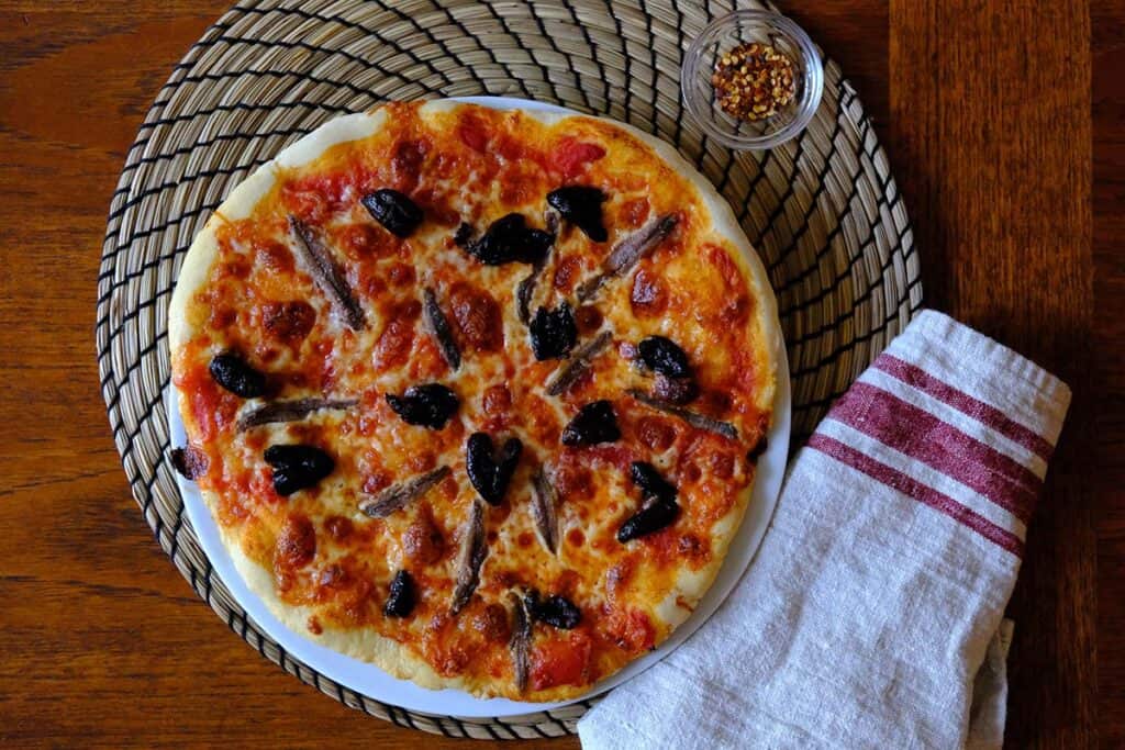 Overhead view of a traditional anchovy pizza topped with briny black olives and fresh mozzarella, served on a white plate.