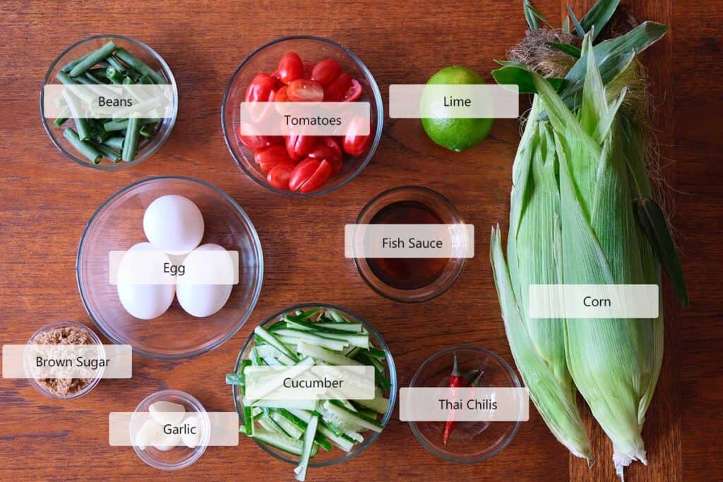 Ingredients for Thai corn salad (Som Tum Khao Pod) on a wooden table, including fresh corn, cherry tomatoes, long green beans, cucumber, eggs, lime, fish sauce, Thai chilies, garlic, and brown sugar.