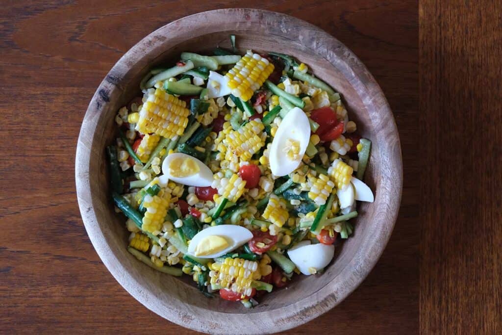 Thai corn salad recipe made with grilled corn, long beans, cherry tomatoes, julienned cucumber, and salted eggs. Served Family style in a large wooden bowl.