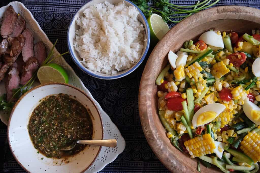 Colorful Thai corn salad with grilled sweet corn, long green beans, cherry tomatoes, cucumber, and salted eggs served in a wooden bowl, paired with sticky rice and grilled moo ping.