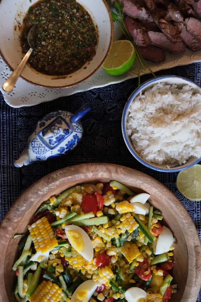 Colorful Thai corn salad with grilled sweet corn, long green beans, cherry tomatoes, cucumber, and salted eggs served in a wooden bowl, paired with rice and grilled pork.