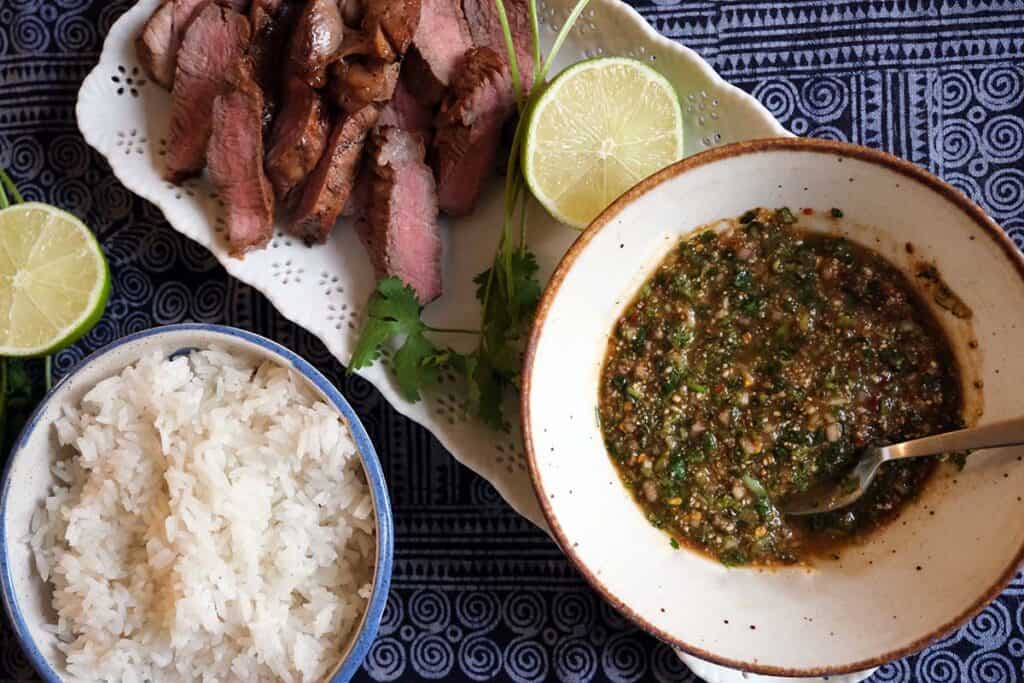 Authentic Thai dipping sauce Nam Jim Jeaw in a bowl alongside sliced grilled steak, sticky rice, and fresh lime wedges.