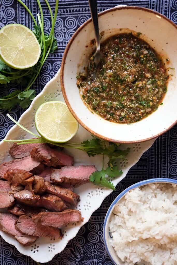 Serving of Nam Jim Jeaw Thai dipping sauce with grilled steak, sticky rice, and lime, showcasing a tangy, spicy, and smoky flavor profile.