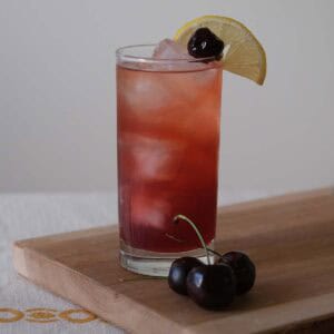 Tall glass of the Cherry Collins Recipe with lemon wedge and Luxardo cherry garnish, showing its jewel-toned purple color over ice.