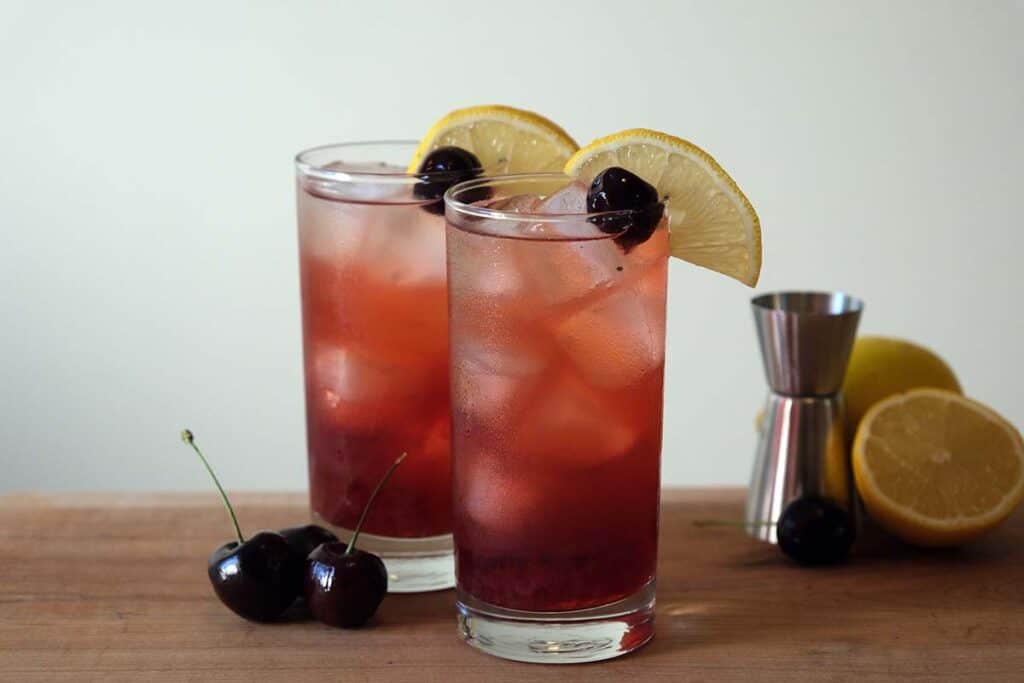 Beautiful purple Cherry Collins Recipe served in tall glasses with ice, lemon slices, and cherries, perfect for gatherings.