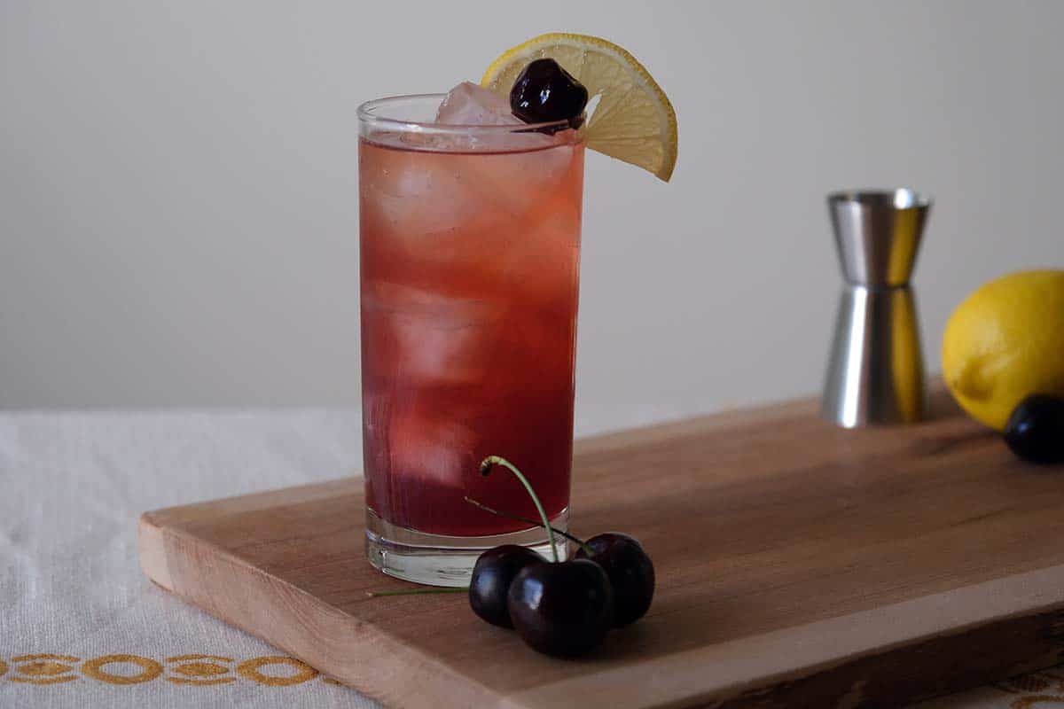 Tall glass of the Cherry Collins Recipe with lemon wedge and Luxardo cherry garnish, showing its jewel-toned purple color over ice.