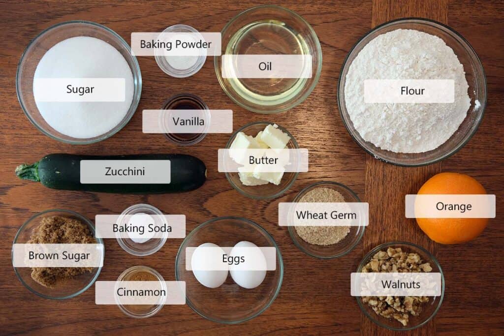 Zucchini and orange muffin recipe ingredients arranged on a wooden tabletop, including fresh zucchini, orange, flour, sugar, eggs, walnuts, and spices.