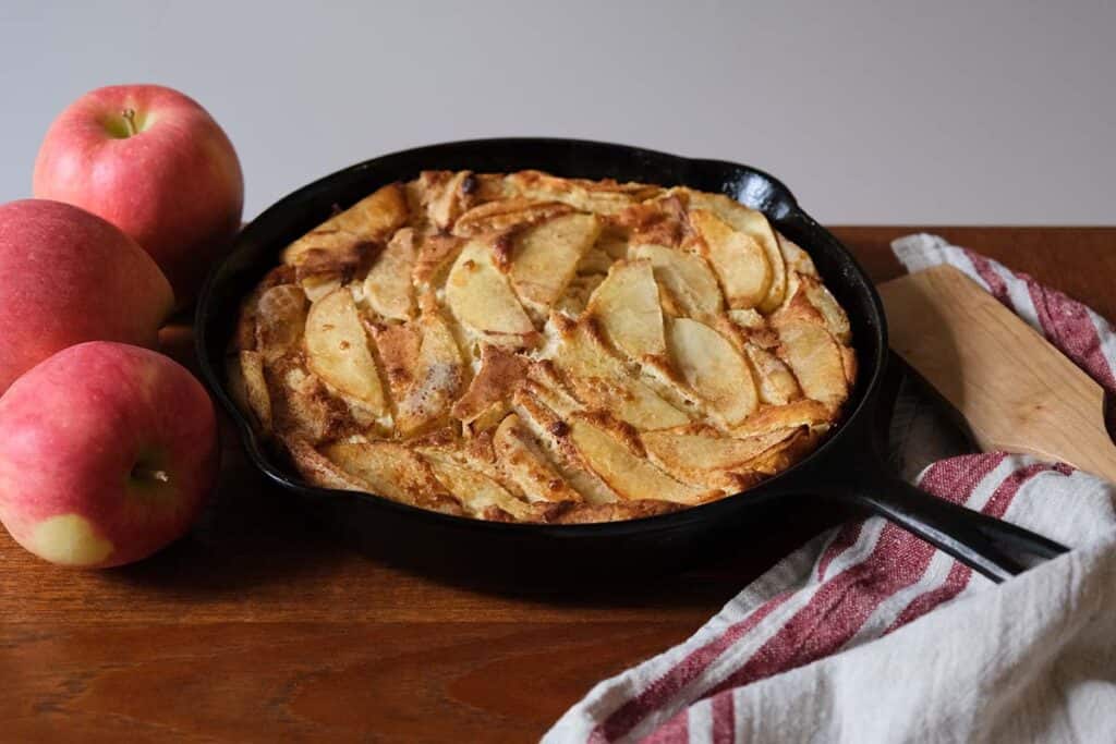 Crisp and golden German Apple Pancake in a black cast iron pan surrounded by fresh apples, perfect for a cozy weekend breakfast.
