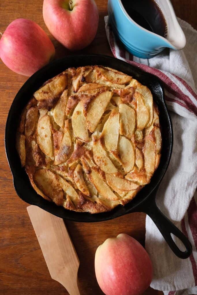 Fluffy German Apple Pancake Recipe in a cast iron skillet surrounded by apples, maple syrup, and a striped tea towel on a wooden table.