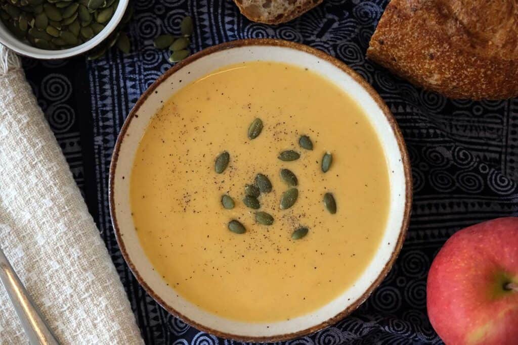 Silky curried apple and butternut squash soup garnished with roasted pumpkin seeds, photographed with bread and fall apples.