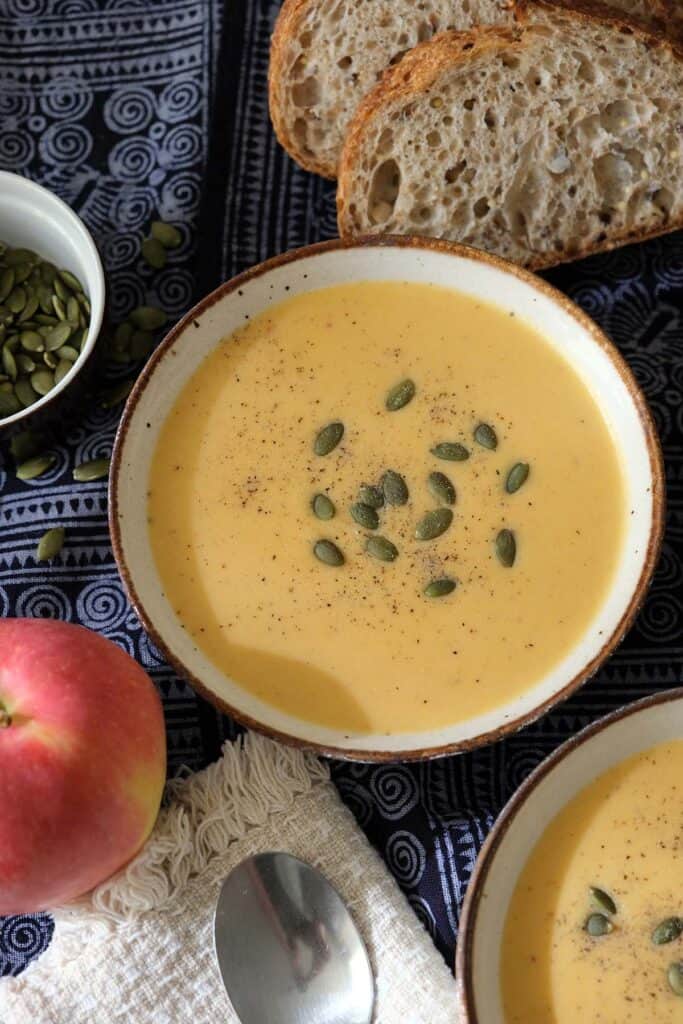 Two bowls of curried apple and butternut squash soup recipe served with crusty bread and roasted pumpkin seeds on a dark patterned cloth.