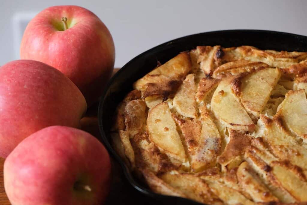 Crisp and golden German Apple Pancake in a black cast iron pan surrounded by fresh apples, perfect for a cozy weekend breakfast.