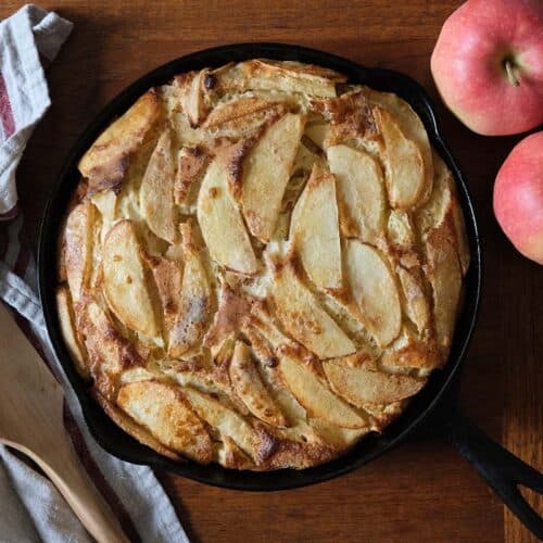 Freshly baked German Apple Pancake in a cast iron skillet with golden apple slices, wooden spatula, and red apples for a farmhouse-style brunch.