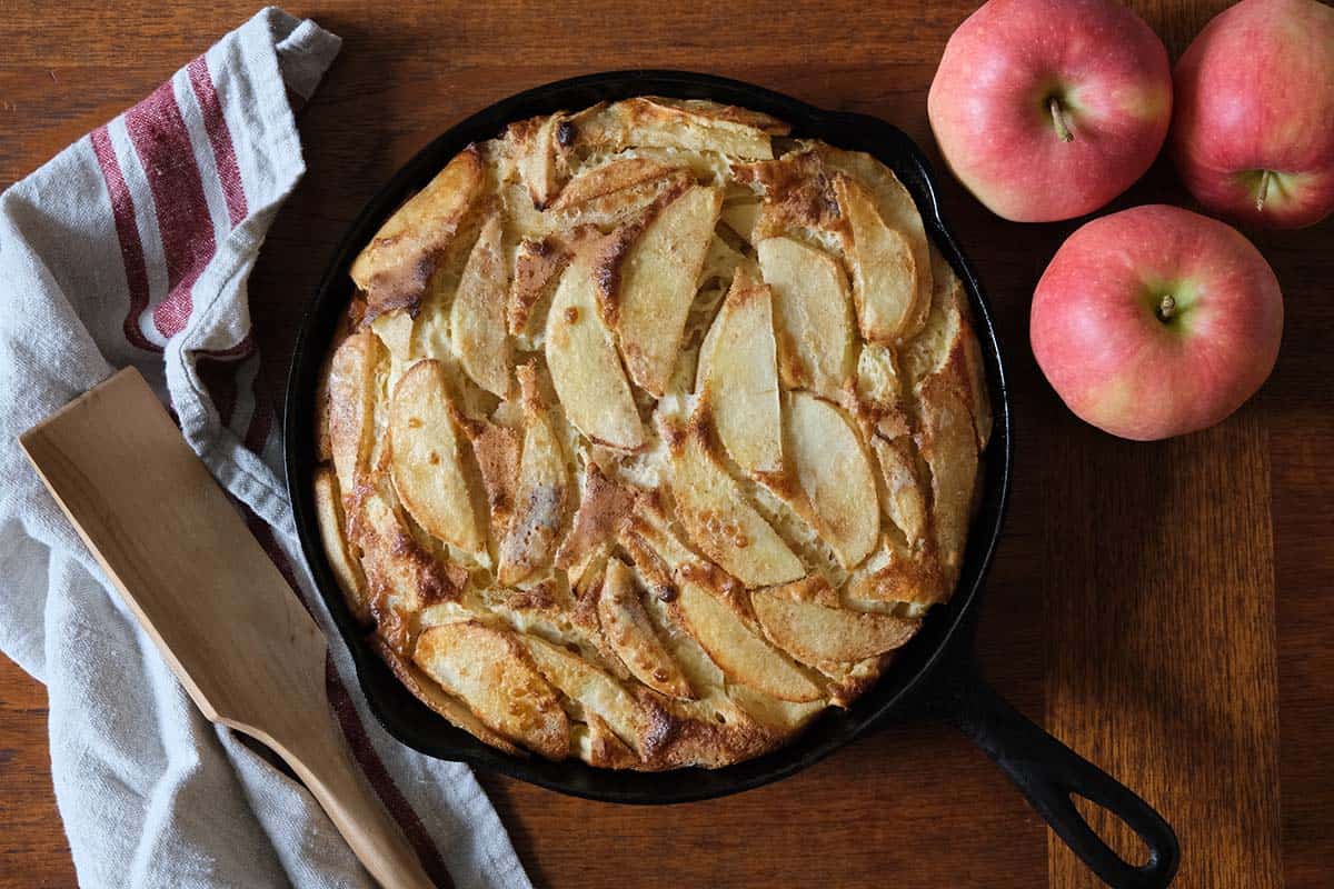 Freshly baked German Apple Pancake in a cast iron skillet with golden apple slices, wooden spatula, and red apples for a farmhouse-style brunch.