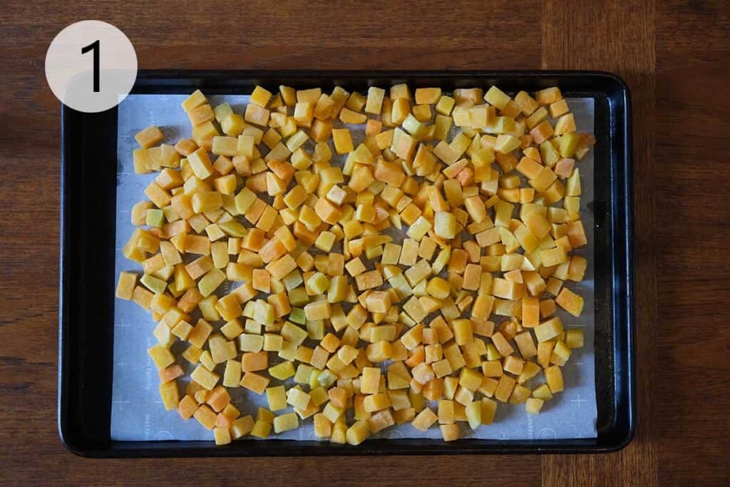 Cubed butternut squash tossed in olive oil and salt on a parchment-lined baking sheet, ready to roast for a silky curried apple and butternut squash soup.