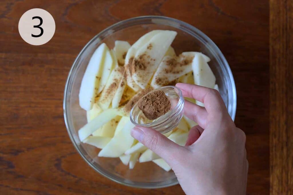Sliced apples tossed in cinnamon and nutmeg in a medium bowl, ready to layer into a warm and fragrant German Apple Pancake.