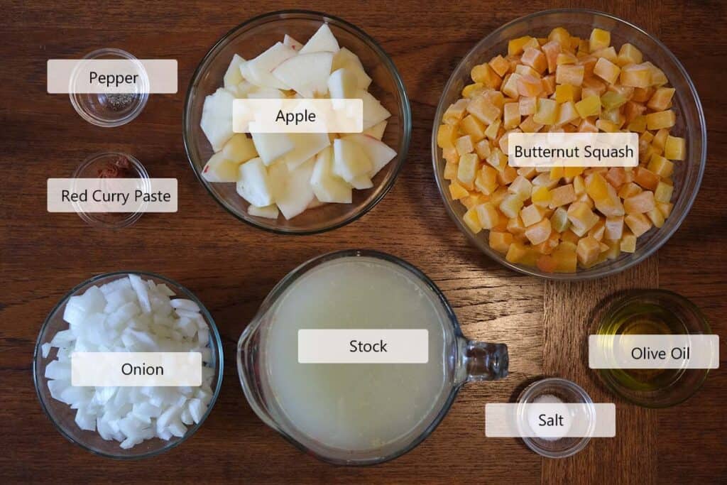 Fresh ingredients for curried apple and butternut squash soup arranged on a wooden surface, including butternut squash, apples, onion, Thai red curry paste, coconut milk, and pumpkin seeds.