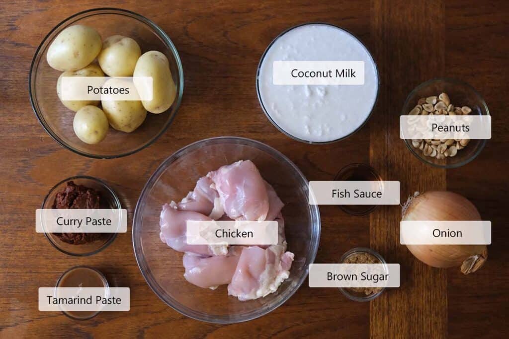Ingredients for Chicken Massaman Curry Recipe including chicken thighs, yellow potatoes, Massaman curry paste, coconut milk, fish sauce, tamarind paste, brown sugar, peanuts, and onions on a wooden table.