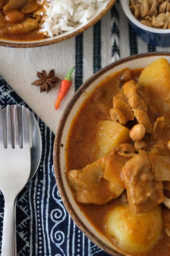 Thai Chicken Massaman Curry Recipe served in bowls with rice, topped with crunchy roasted peanuts, surrounded by a blue napkin and aromatic spices.