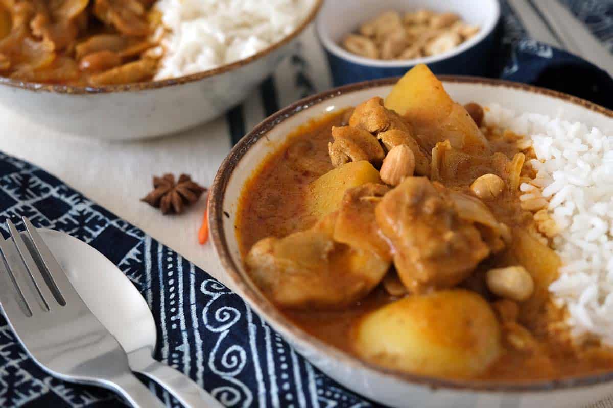 Creamy Chicken Massaman Curry with tender chicken, buttery potatoes, and roasted peanuts served over jasmine rice in bowls, styled with a blue napkin and spices.