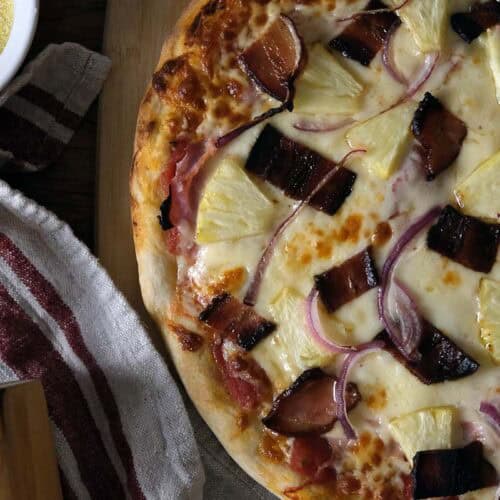 Homemade Hawaiian pizza with melted cheese and caramelized toppings, styled on a wooden board with a rolling pin and bowl of cornmeal.