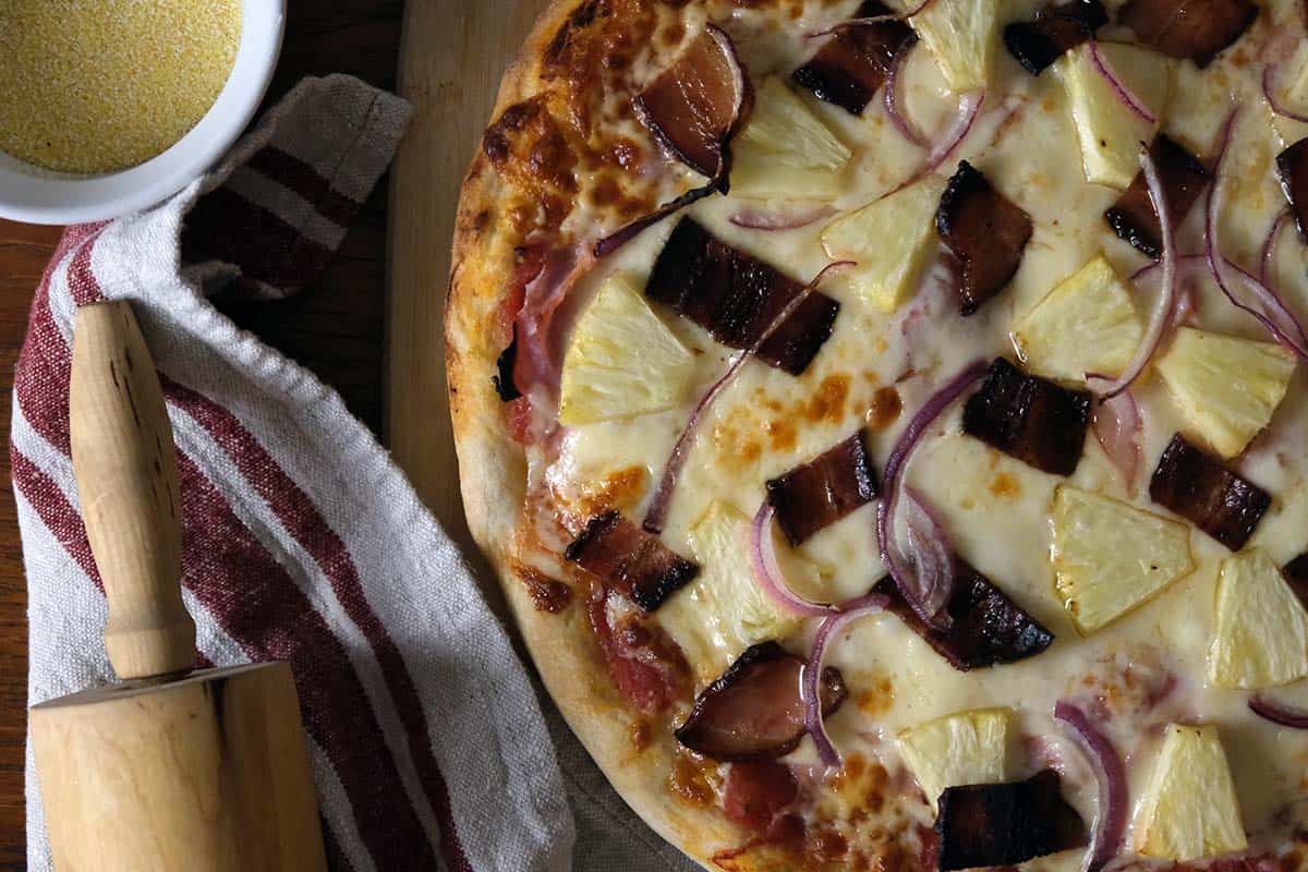 Homemade Hawaiian pizza with melted cheese and caramelized toppings, styled on a wooden board with a rolling pin and bowl of cornmeal.