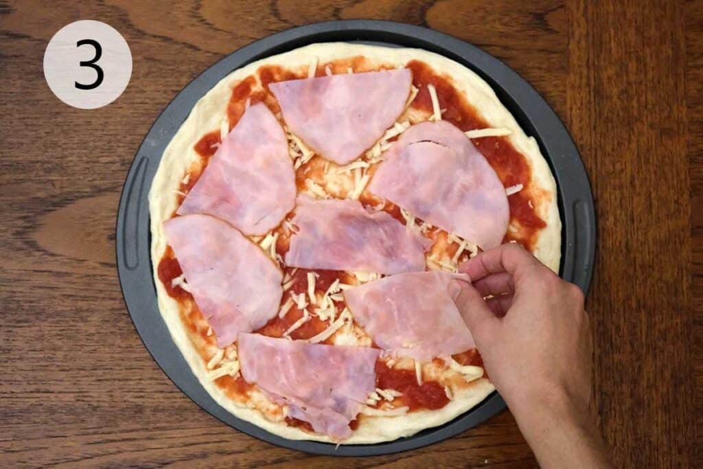 Layering sliced ham over a base of mozzarella and sauce while assembling a homemade Hawaiian pizza.