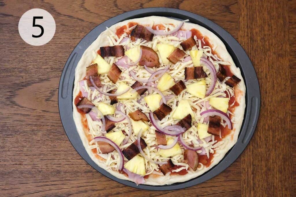 Topping the pizza with pineapple chunks, cooked bacon, and red onion for a classic Hawaiian pizza before baking.