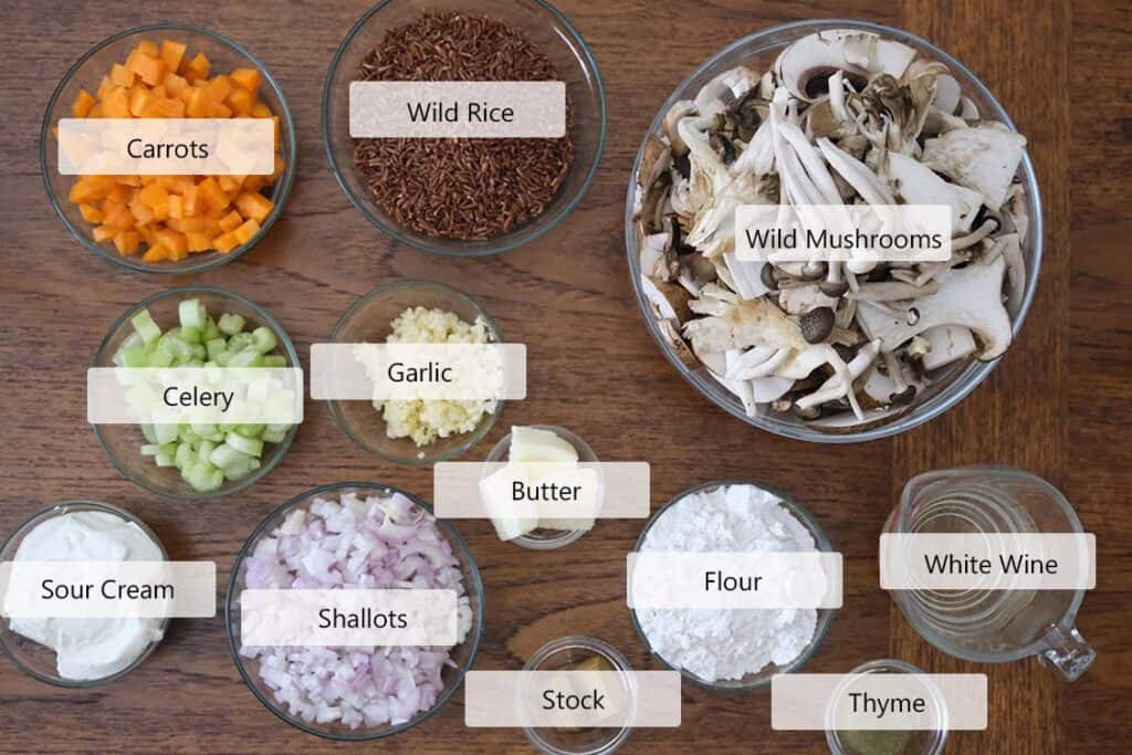 Overhead view of fresh wild mushrooms, shallots, carrots, celery, thyme, wild rice, and sour cream arranged for a creamy wild mushroom soup recipe.