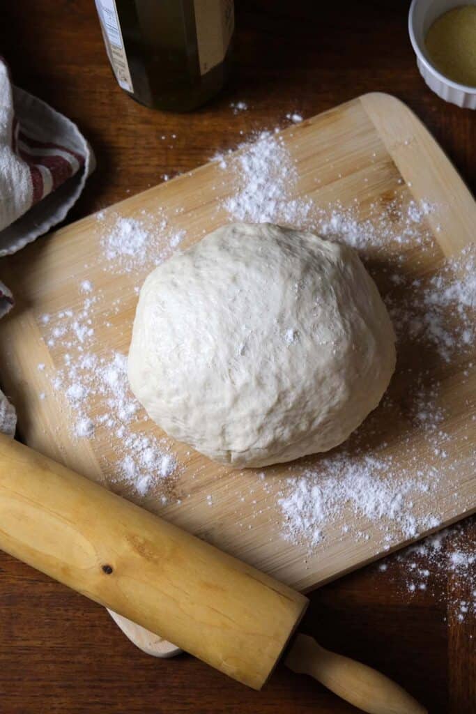 Top view of a ball of thin crust pizza dough on a floured board with a rolling pin and kitchen towel, ready to shape for pizza.