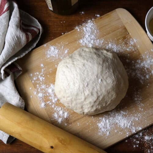 Preparing thin crust pizza dough for homemade pizza, showing a smooth dough ball on a floured surface with a rolling pin nearby.