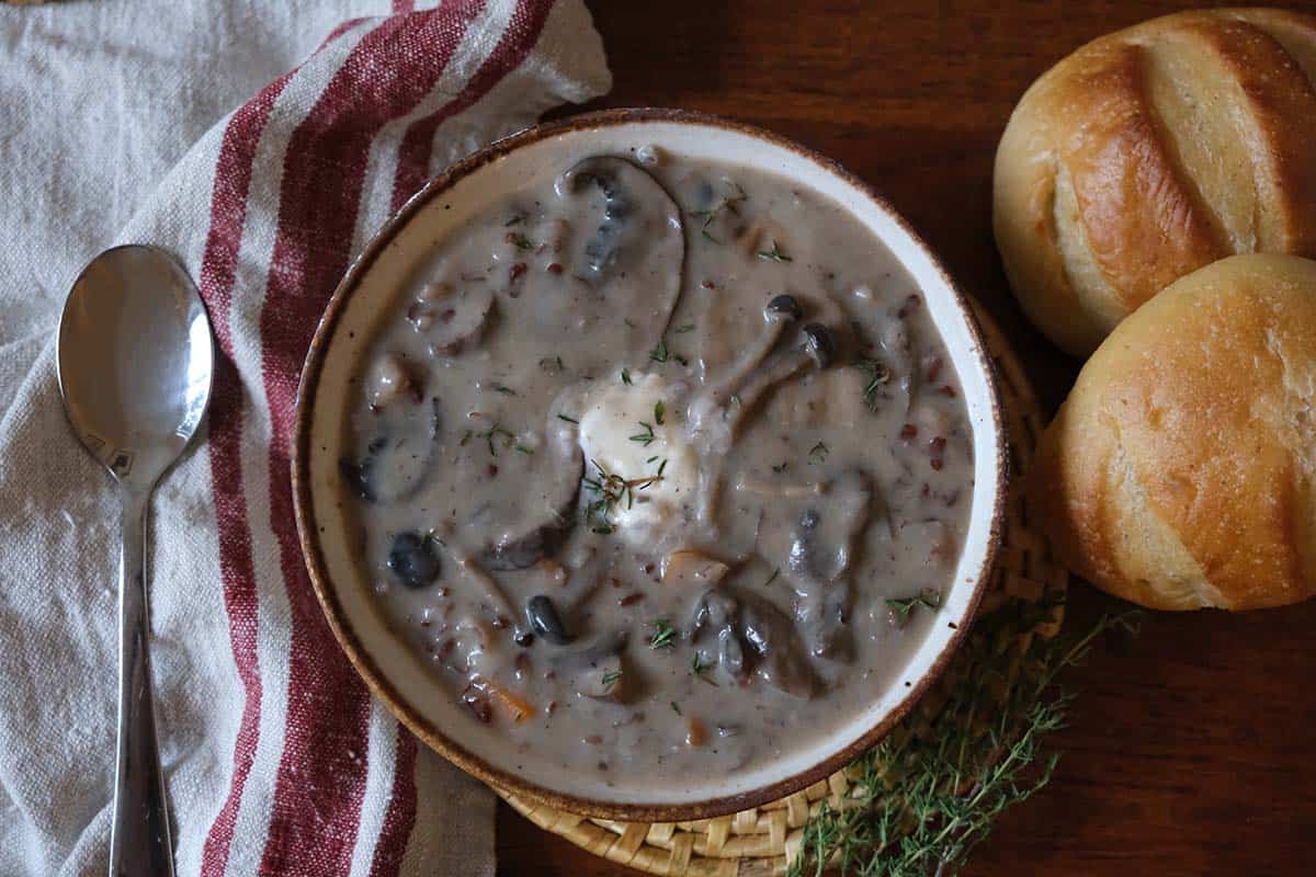 Rustic presentation of creamy wild mushroom soup recipe beside golden dinner rolls and a red striped linen napkin.