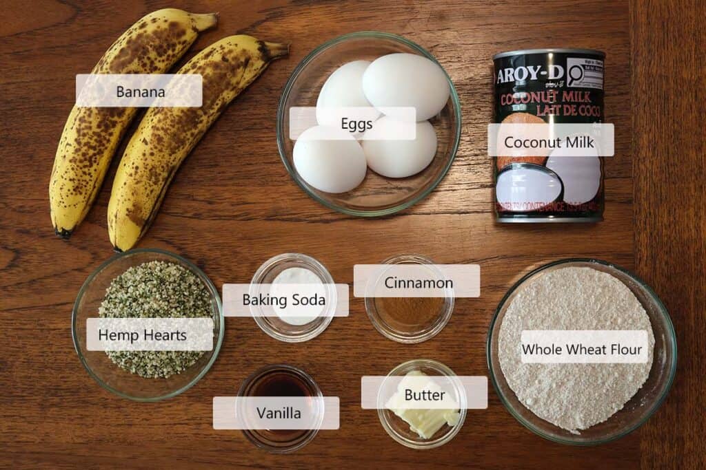 Ingredients for whole wheat banana pancakes including ripe bananas, whole wheat flour, eggs, coconut milk, hemp hearts, cinnamon, and baking soda arranged on a countertop.