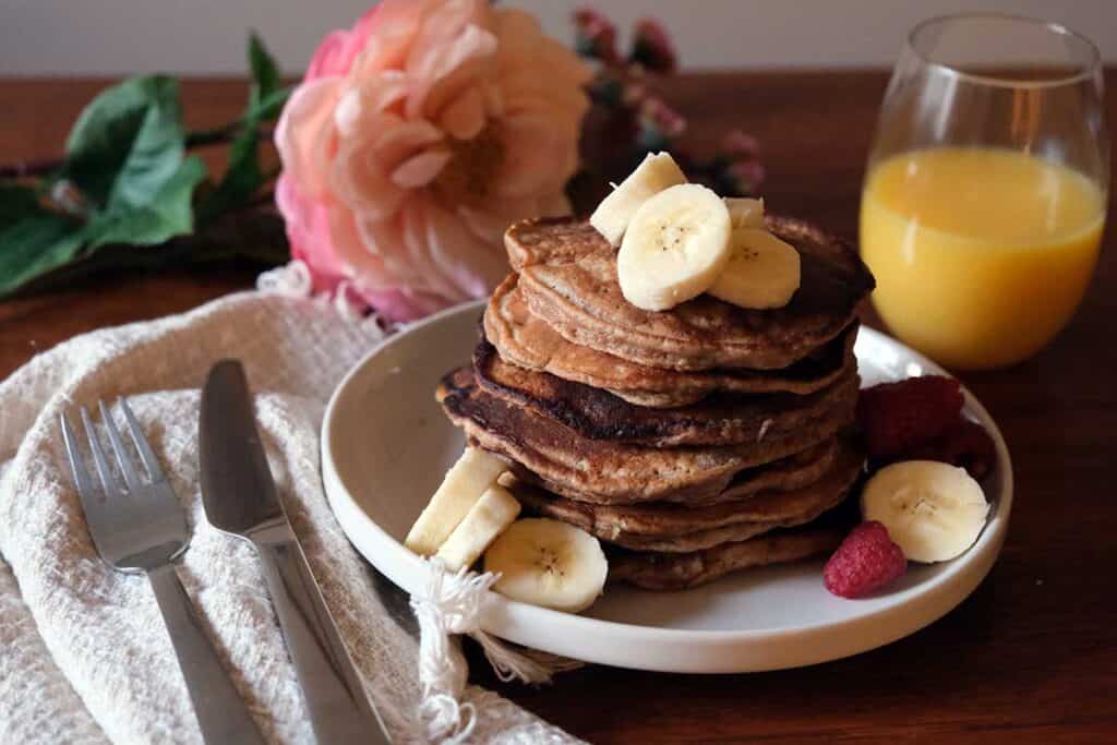 Fluffy whole wheat banana pancakes with fresh berries and sliced bananas drizzled in maple syrup, styled for Sunday brunch with orange juice and flowers.